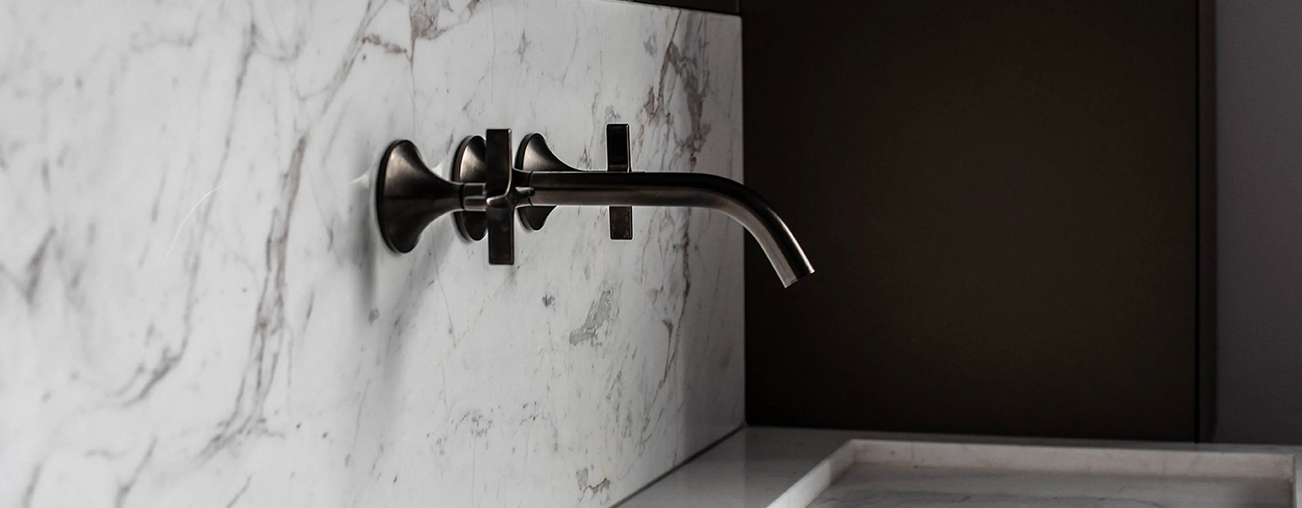 Wandmontierte, dunkle Waschtisch-Armatur mit gebogenem Auslauf und Kreuzgriffen vor einer eleganten Marmorwand in einem stilvollen, modernen Badezimmer.