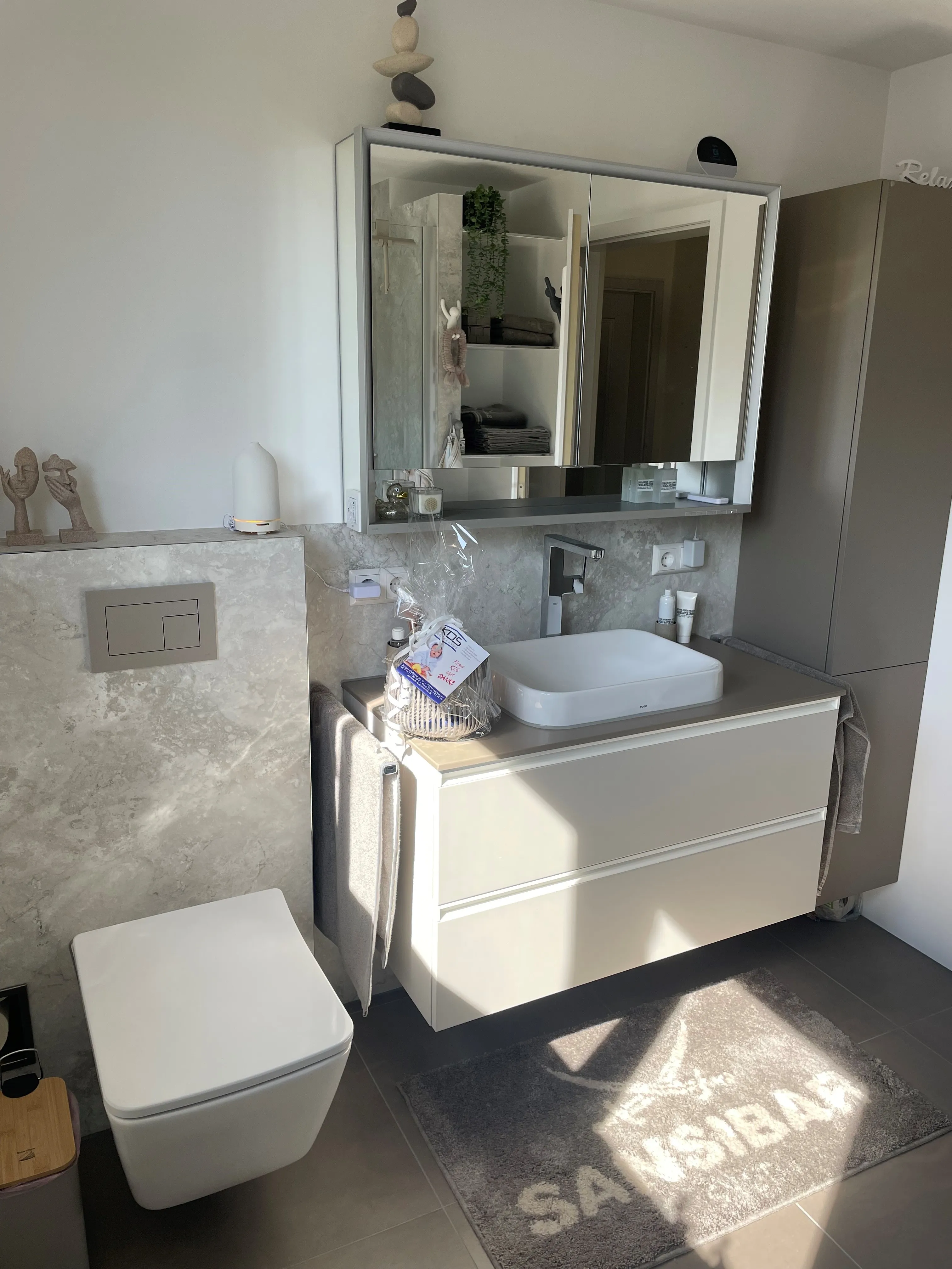 Modernes Badezimmer mit wandhängendem WC, hellem Waschtisch, großem Spiegelschrank und Steinoptik-Rückwand.