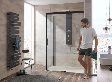 Mann steht in modernem Bad vor einer Glasduschkabine  HSK Atelier Plan mit schwarzem Rahmen, grauen Fliesen und Holzfußboden. Ein schwarzer Handtuchheizkörper ist an der Wand.