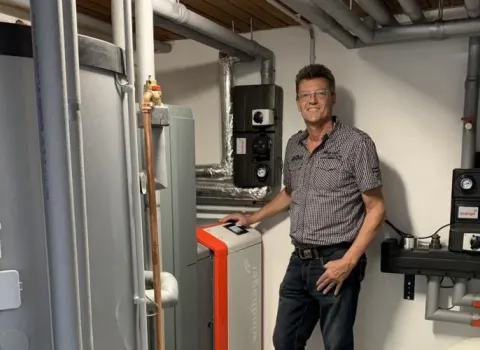 Mann steht in Heizungsraum neben moderner Heizungsanlage mit Pufferspeicher und Rohrleitungen.