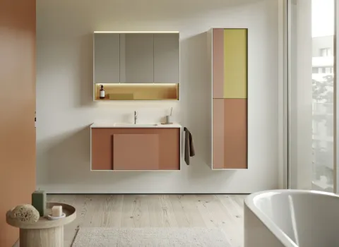 Ein Badezimmer im gerahmten Look zeigt Hängemöbel mit klar definierten weißen Rahmen, die farblich abgesetzte Fronten in Rostrot, Beige und Senfgelb einrahmen. Der Waschtisch und der hohe Seitenschrank wirken durch die Rahmenstruktur besonders strukturiert und hochwertig.