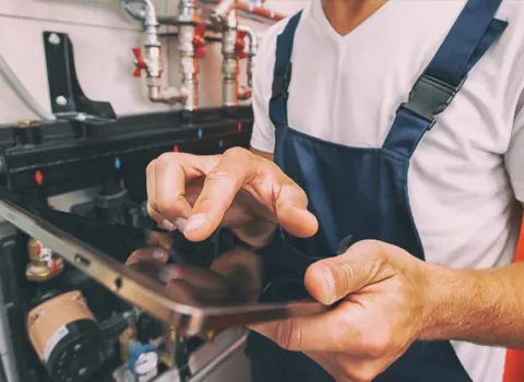 Anlagemechaniker, der Firma KDS Haustechnische Anlagen GmbH, in blauer Latzhose bedient ein Tablet vor einer Heizungsinstallationsanlage.