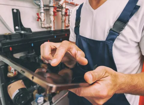 Anlagemechaniker, der Firma KDS Haustechnische Anlagen GmbH, in blauer Latzhose bedient ein Tablet vor einer Heizungsinstallationsanlage.