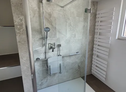 Große, bodengleiche Dusche mit Regenduschkopf, Haltegriff und Duschsitz vor steinfarbener Wand.