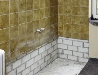 Unrenovierter Badezimmerbereich mit braunen Wandfliesen aus den 1970er-Jahren. Der ehemalige Badewannenplatz ist freigelegt: Die unteren Fliesen wurden entfernt und durch eine halbhoch gemauerte Wand aus weißen Kalksandsteinen ersetzt.