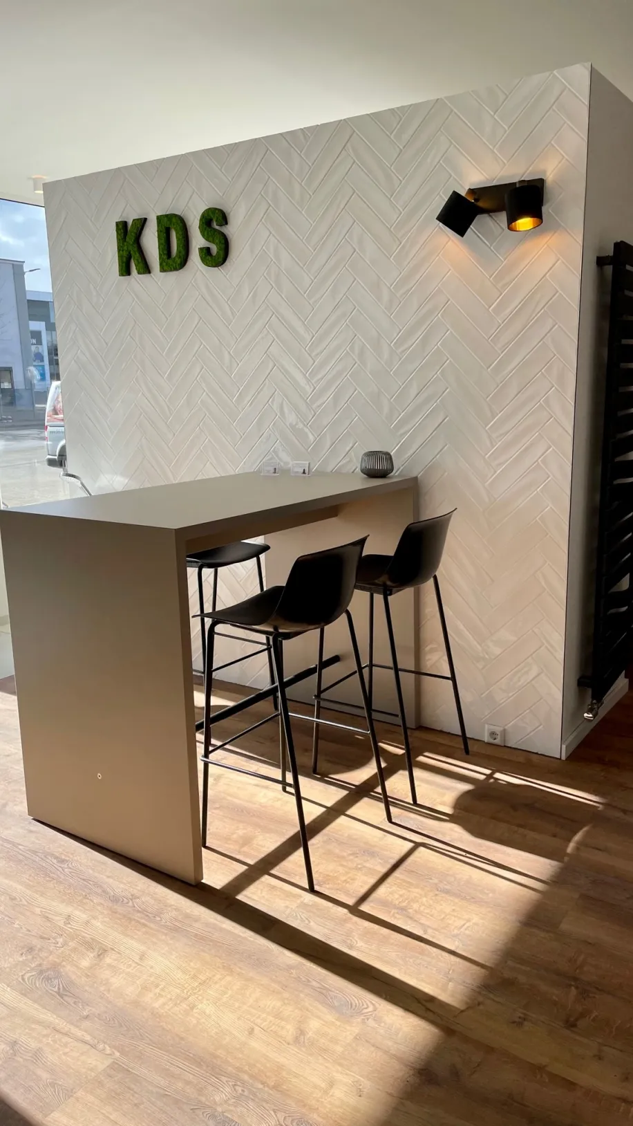 Ein heller Ausstellungsbereich mit modernem Bartisch und zwei schwarzen Barhockern steht vor einer weiß gefliesten Wand im Fischgrätmuster.