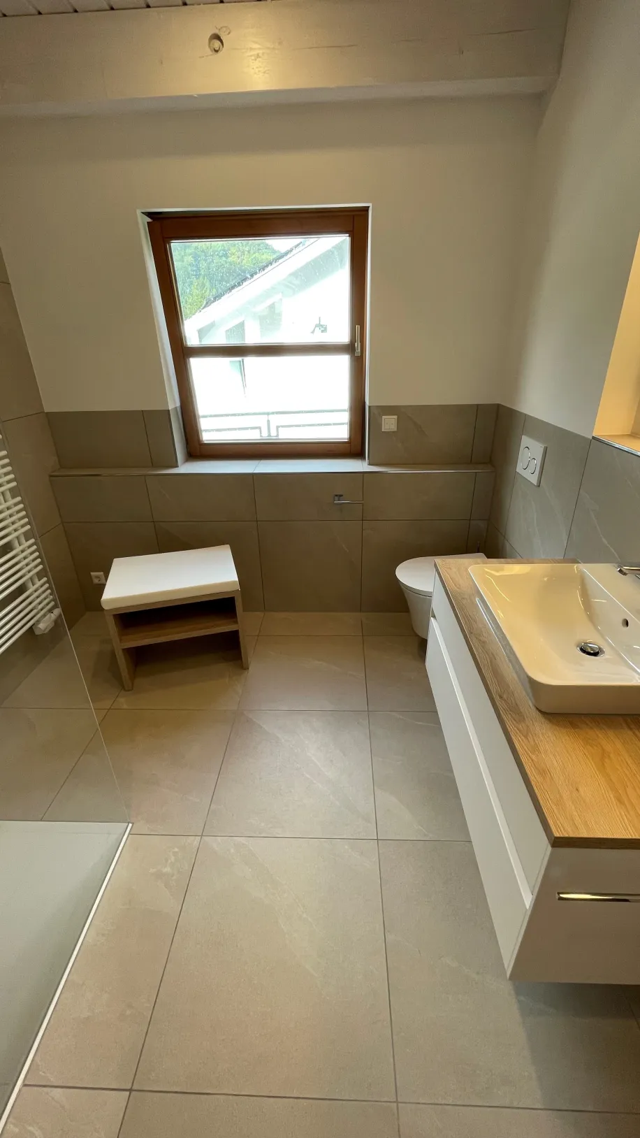 Ein helles, natürlich gestaltetes Badezimmer mit großformatigen beigen Bodenfliesen und Holz-Akzenten.