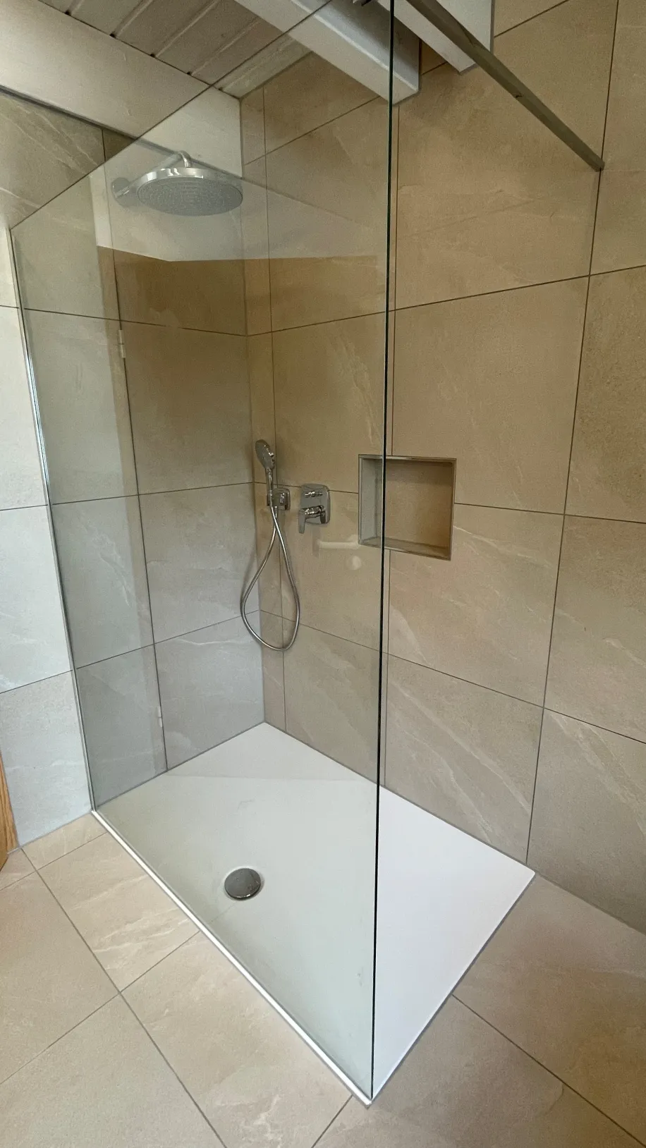 Eine moderne, bodengleiche Dusche mit großer Glasabtrennung ist in einem Badezimmer mit beigen Natursteinfliesen zu sehen. 