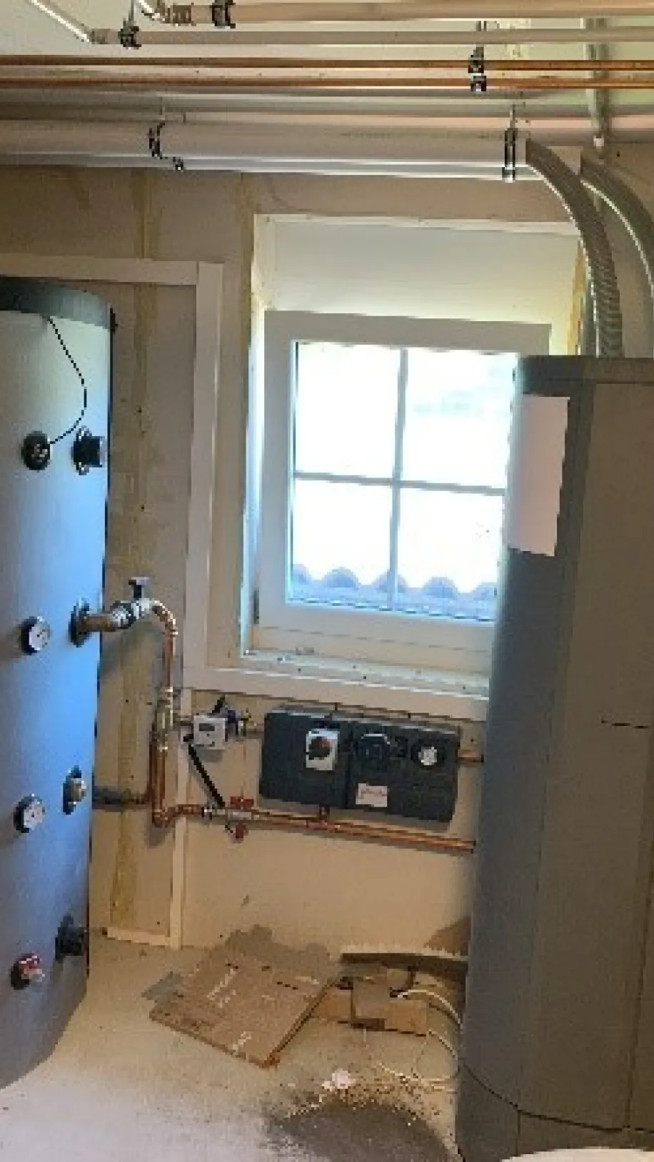 Heizungsraum mit moderner Windhager-Pelletheizung und zwei großen Pufferspeichern, installiert mit Kupferrohren und Lüftungsleitung unter dem Fenster.