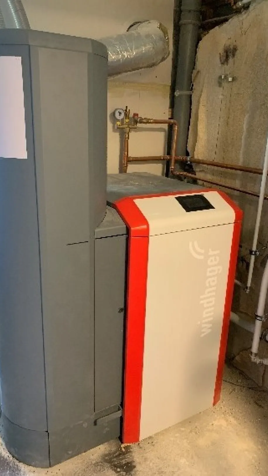 Windhager-Heizungsanlage in einem Heizraum installiert, mit angeschlossenen Kupferrohren und Lüftungsrohr, moderne und energieeffiziente Technik.
