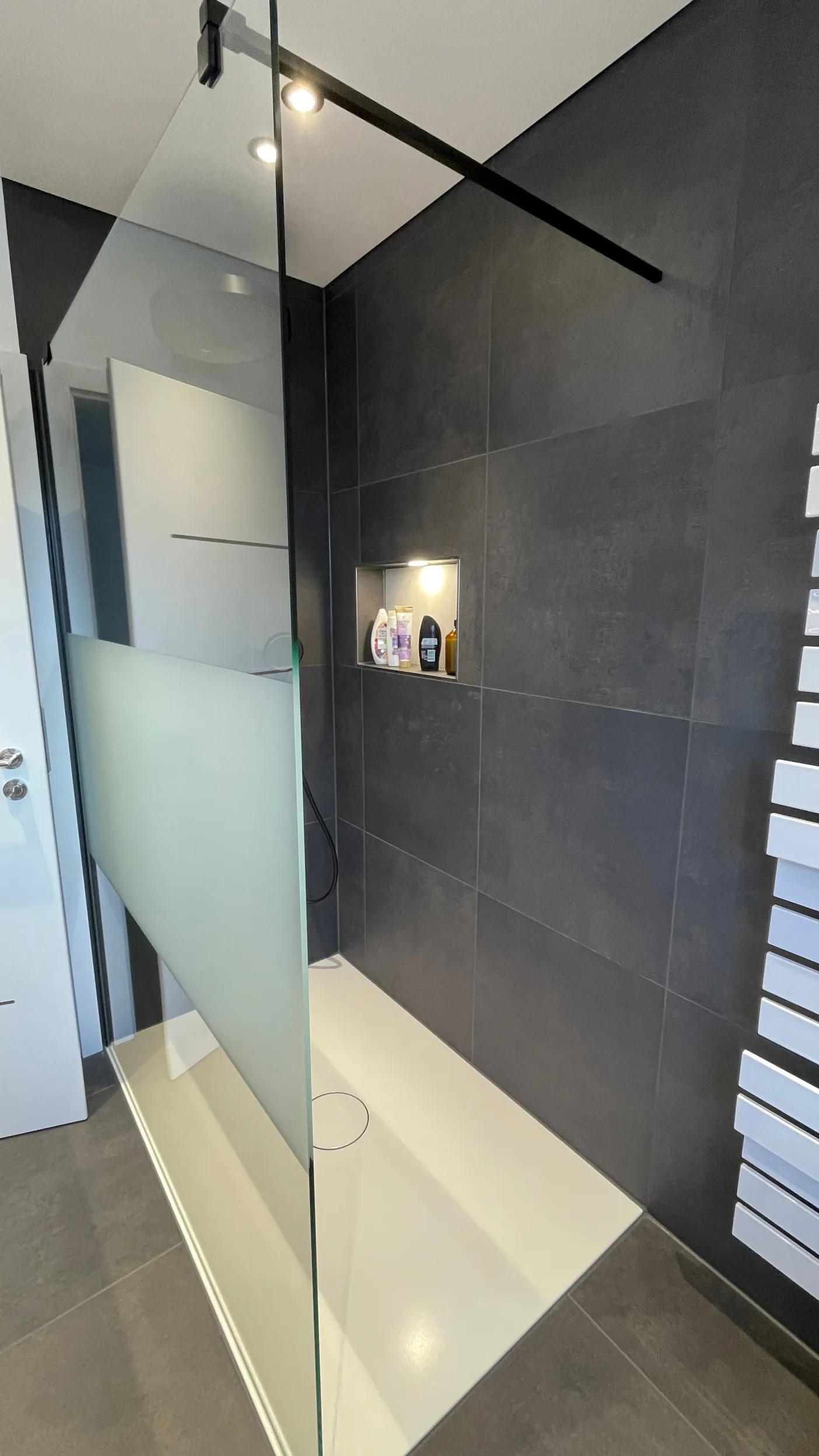 Eine moderne, begehbare Dusche mit dunklen Wandfliesen und einer großen, rahmenlosen Glaswand mit satinierter Fläche zur Sichtbegrenzung. 