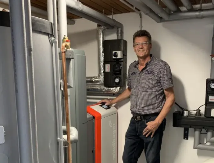 Mann steht in Heizungsraum neben moderner Heizungsanlage mit Pufferspeicher und Rohrleitungen.