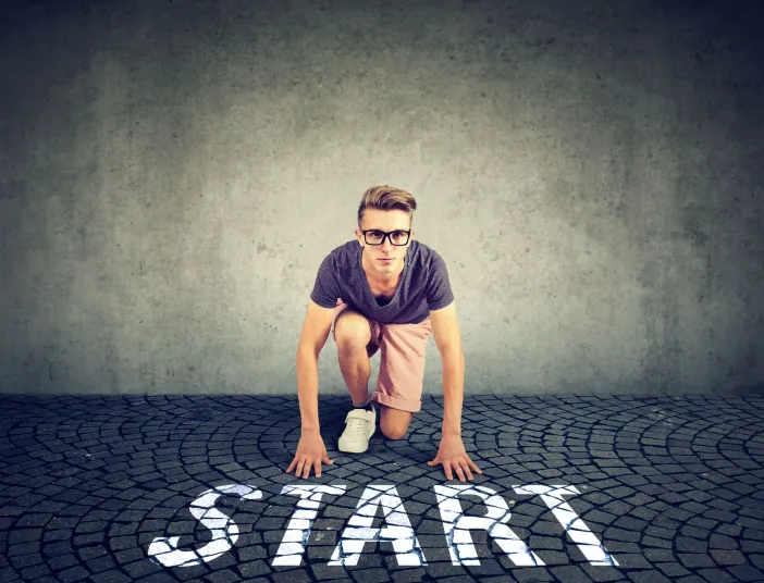 Vor einer schlichten grauen Wand geht ein junger Mann mit Brille in Startposition auf einem Kopfsteinpflasterboden.