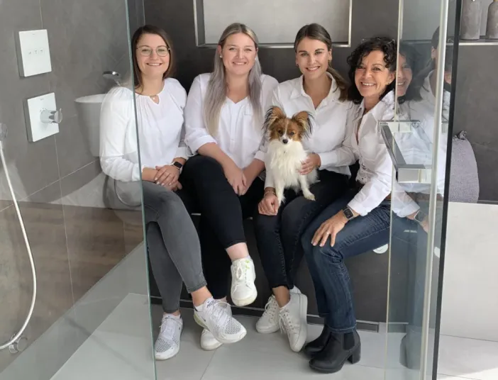 Das vierköpfige Büroteam von KDS-Haustechnik posiert sitzend mit einem Schoßhündchen in der Badausstellung.