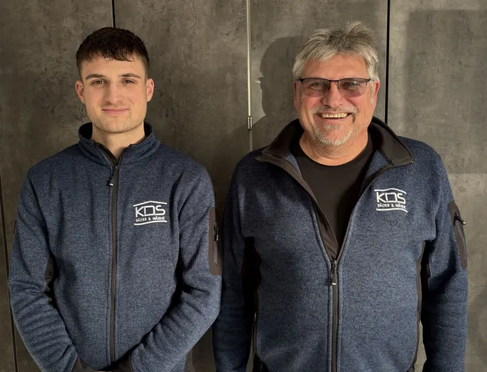 Zwei Männer des Heizungsteams von KDS-Haustechnik stehen vor eine dunklen Wand und schauen in die Kamera.
